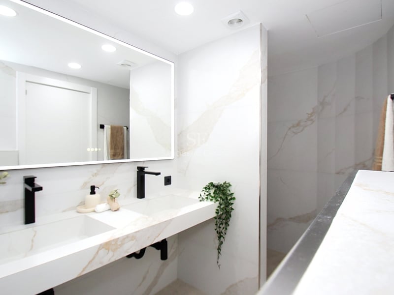 Ljust badrum med dubbelt tvättställ i vit marmor, mattsvarta kranar och en stor bakgrundsbelyst spegel.