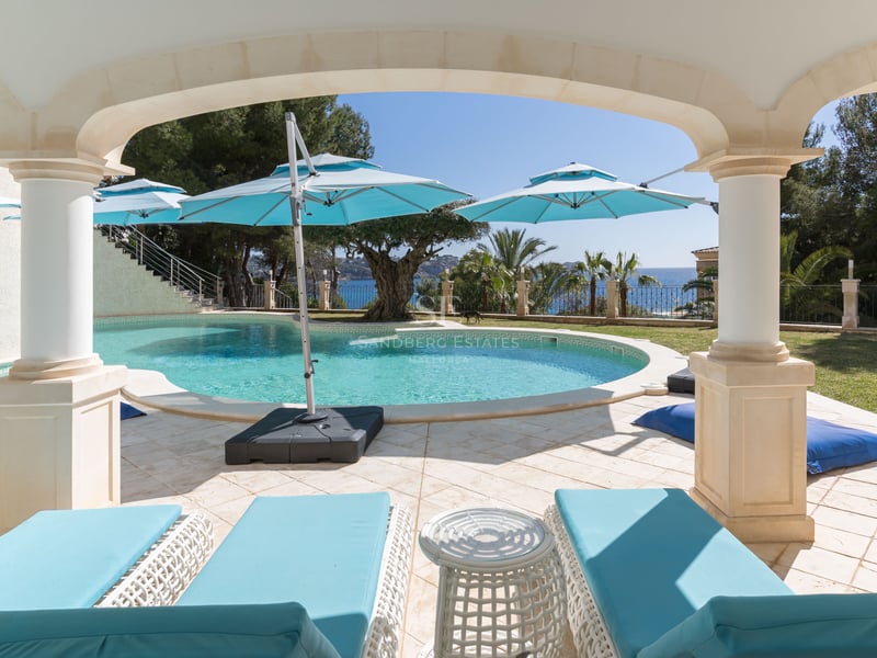 Ein geschwungener türkisfarbener Swimmingpool, umgeben von Natursteinfliesen und türkisfarbenen Liegen unter einem Torbogen.