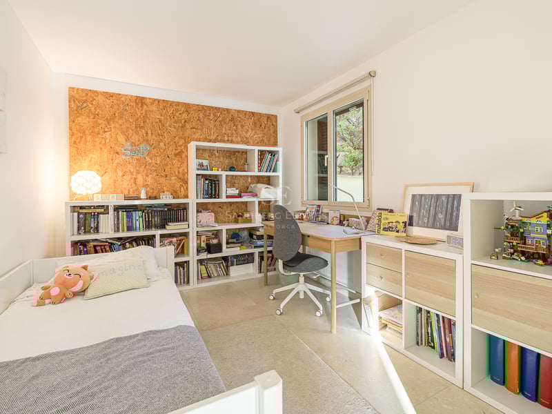 Barnrum med vit säng, skrivbord i trä och bokhyllor fyllda med böcker under ett ljust fönster.