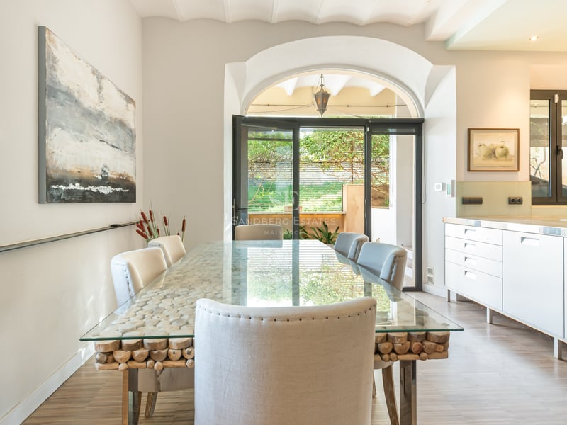 Ljus matsal med ett glasbord på en bas av trästockar, vita stolar och ett valv mot uteplatsen.