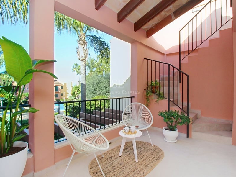 Laxrosa övertäckt terrass med vita Acapulco-stolar, träbjälkar och utsikt över poolområdet.