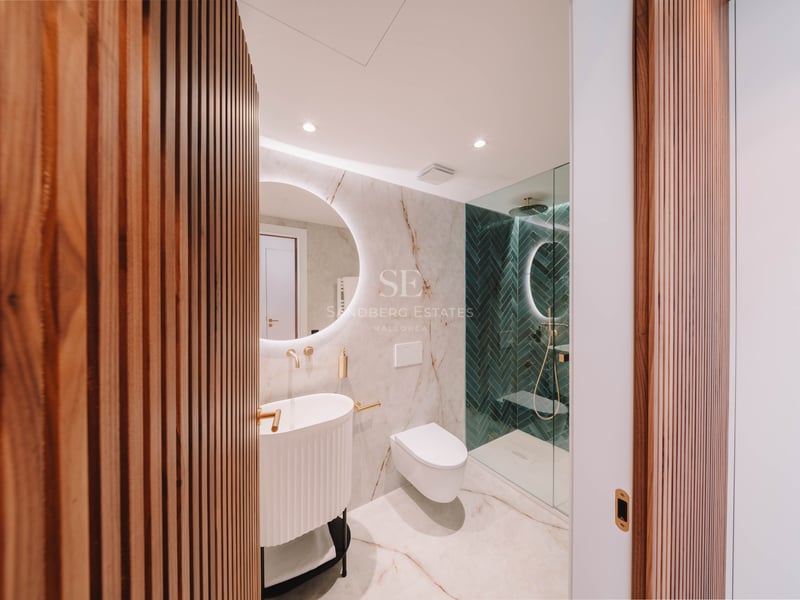 Salle de bain moderne avec marbre, miroir rond rétroéclairé, robinetterie dorée et douche en chevrons verts.