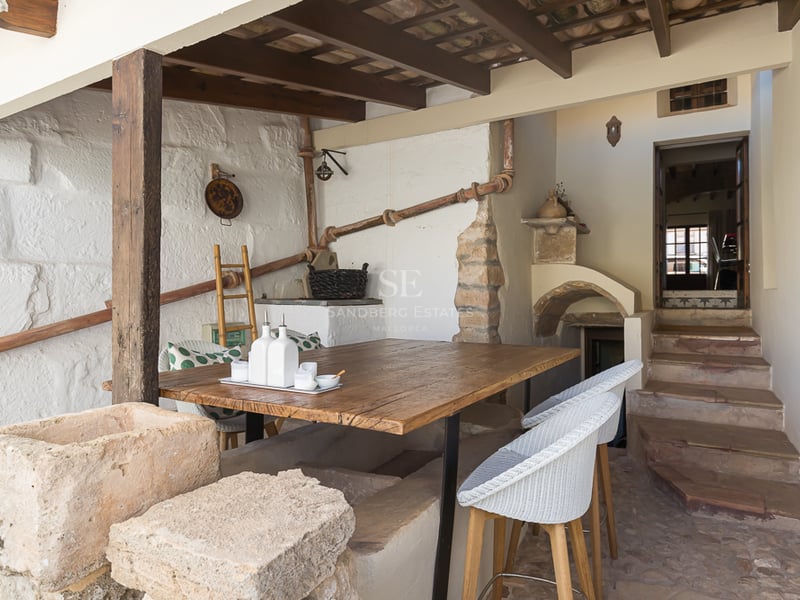 Coin repas extérieur rustique avec table en bois, murs en pierre, poutres apparentes et escaliers en pierre.