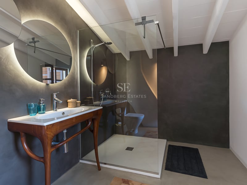 Salle de bain moderne avec murs en microciment gris foncé, miroir circulaire rétroéclairé et meuble en bois sculpté.