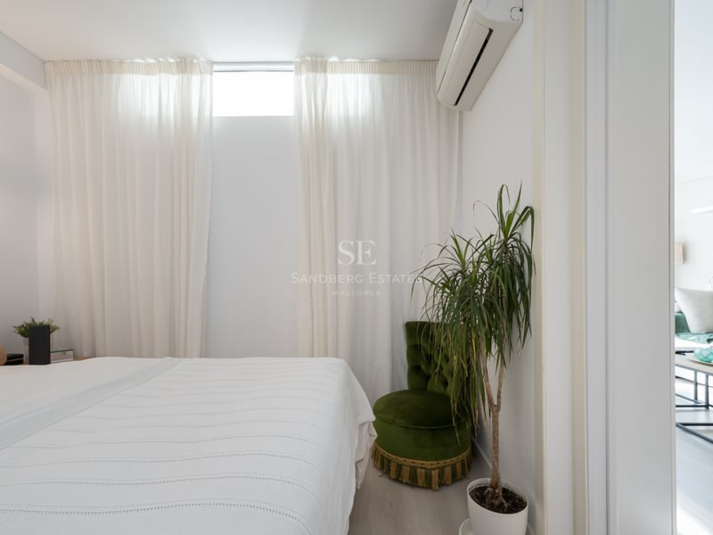 Clean white bedroom featuring a bed, green velvet chair, indoor plant, and a doorway looking into the living room.