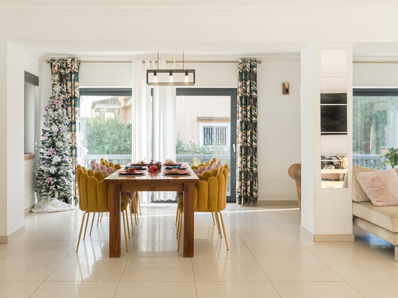 Espace salle à manger moderne avec table en bois, chaises en velours jaune, sapin de Noël et grandes baies vitrées.