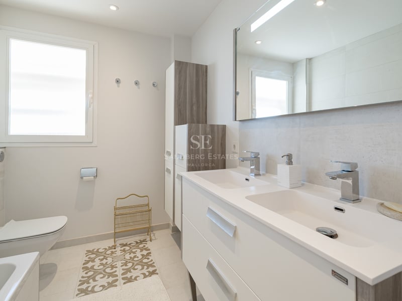 Bright modern bathroom with a double white vanity, large mirror, wall-hung toilet, and natural window light.