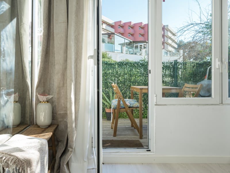 View through a glass door to a wooden terrace with outdoor furniture and a privacy hedge.
