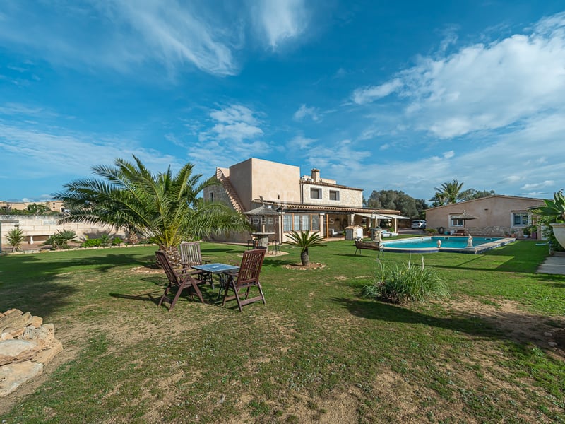 Extérieur d'une villa rustique avec pelouse, palmiers, piscine privée et mobilier de jardin sous un ciel bleu.