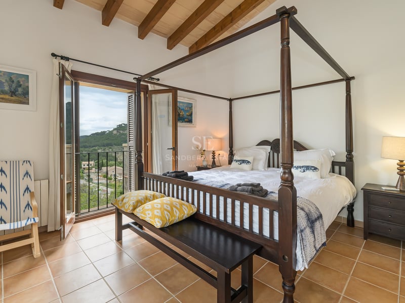 Chambre principale avec lit à baldaquin, poutres en bois, sol en terre cuite et balcon donnant sur les collines.