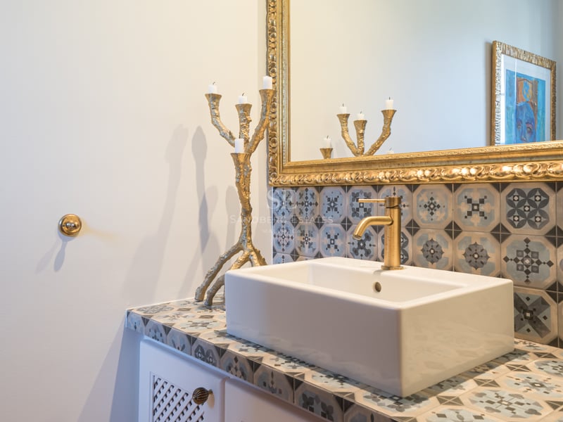 Close-up of a white vessel sink on a patterned tile countertop with a gold faucet and ornate gilded mirror.