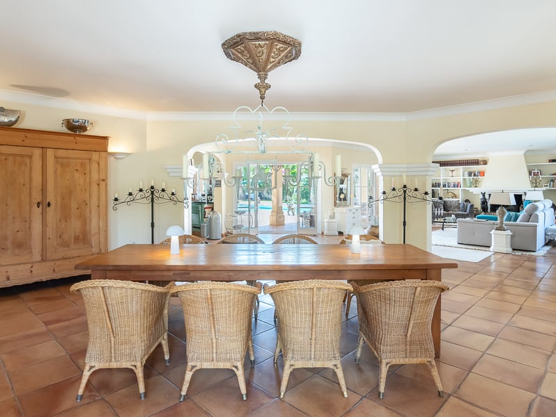 Salle à manger spacieuse avec table en bois, chaises en osier, sol en terre cuite et vues sur la piscine.