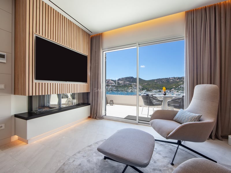 Salon contemporain avec fauteuil, cheminée, télévision sur mur en bois et vue panoramique sur le port depuis le balcon.