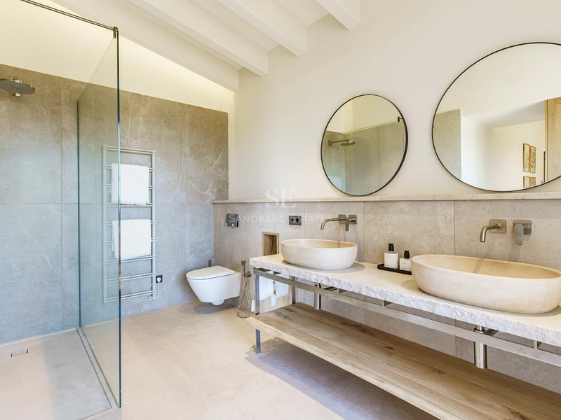 Luxuriöses Badezimmer mit zwei Steinwaschbecken auf Marmorplatte, Rundspiegeln und begehbarer Glasdusche.