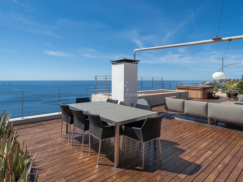 Modern rooftop terrace with wooden decking, dining table, lounge area, and glass railings overlooking the blue sea.