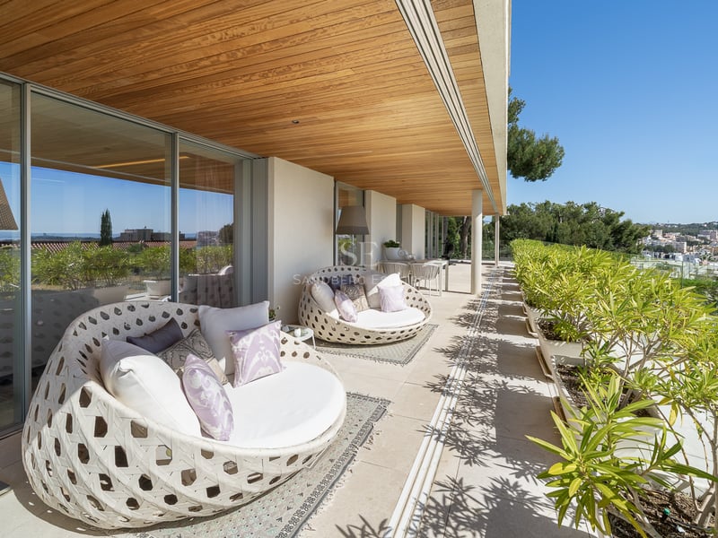 Sonnige Terrasse mit runden weißen Loungesesseln, Holzdecke und Blick auf die mediterrane Bepflanzung.
