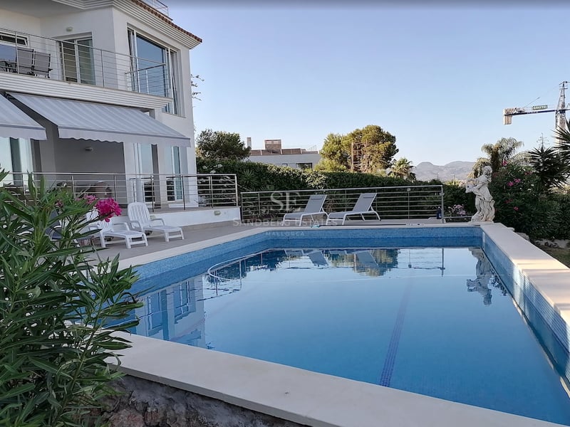 Une piscine rectangulaire en mosaïque bleue devant une villa blanche avec des chaises longues et des plantes.