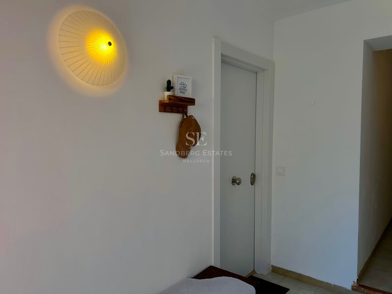 A white hallway featuring a round pleated wall lamp, a small wooden shelf with a hat, and a white door.