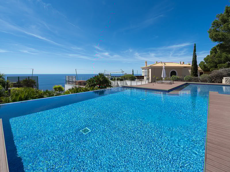 Large infinity swimming pool with blue tiles overlooking the sea and a stone terrace under a clear sky.