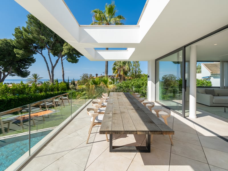 Terrasse blanche moderne avec grande table en bois, garde-corps en verre et vue sur la mer et la piscine.