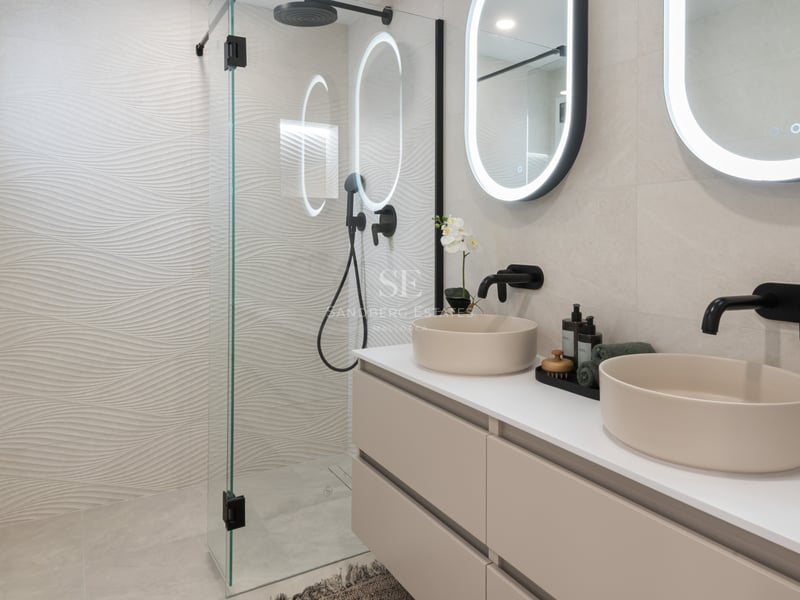 Salle de bain moderne avec deux vasques beiges, miroirs rétroéclairés et douche avec carrelage ondulé.