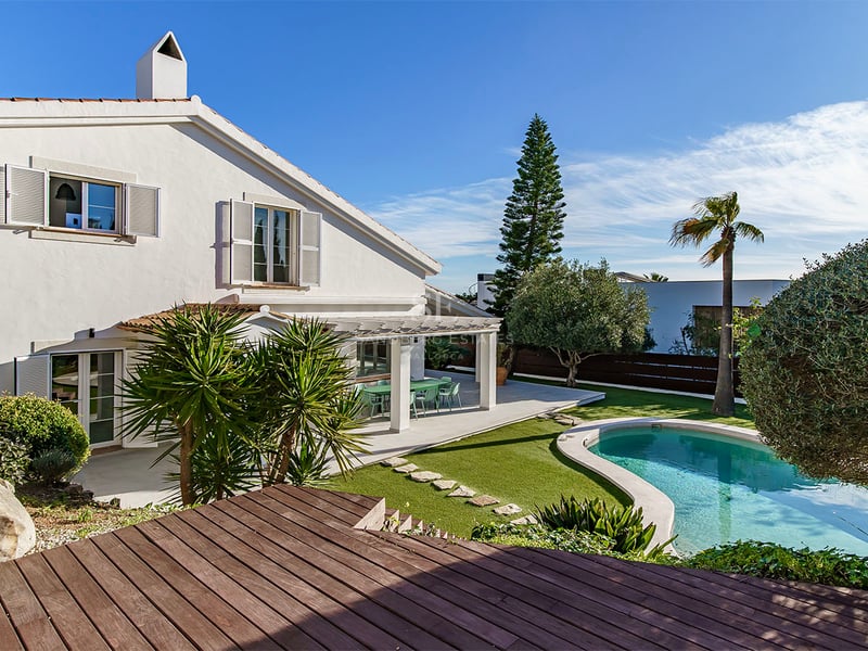Vit tvåvåningsvilla med en njurformad pool, trädäck och välskött trädgård under klarblå himmel.