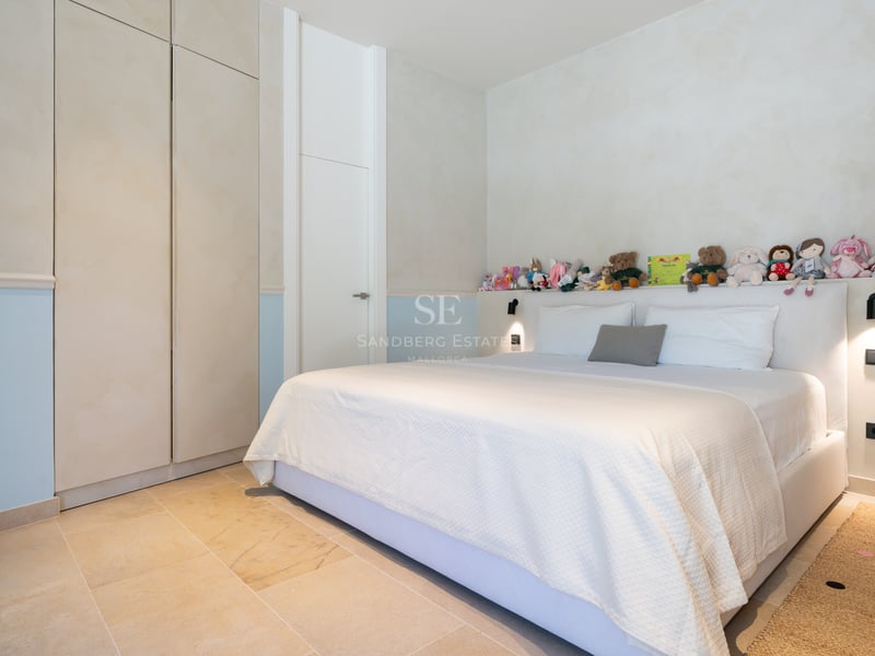 Chambre lumineuse avec un grand lit, linge de lit blanc, sol en pierre naturelle et placards intégrés crème.