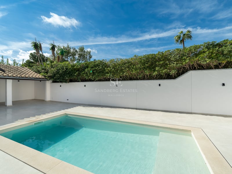 Piscine turquoise sur terrasse blanche avec mur d'intimité, haies vertes et cuisine d'été sous ciel bleu.
