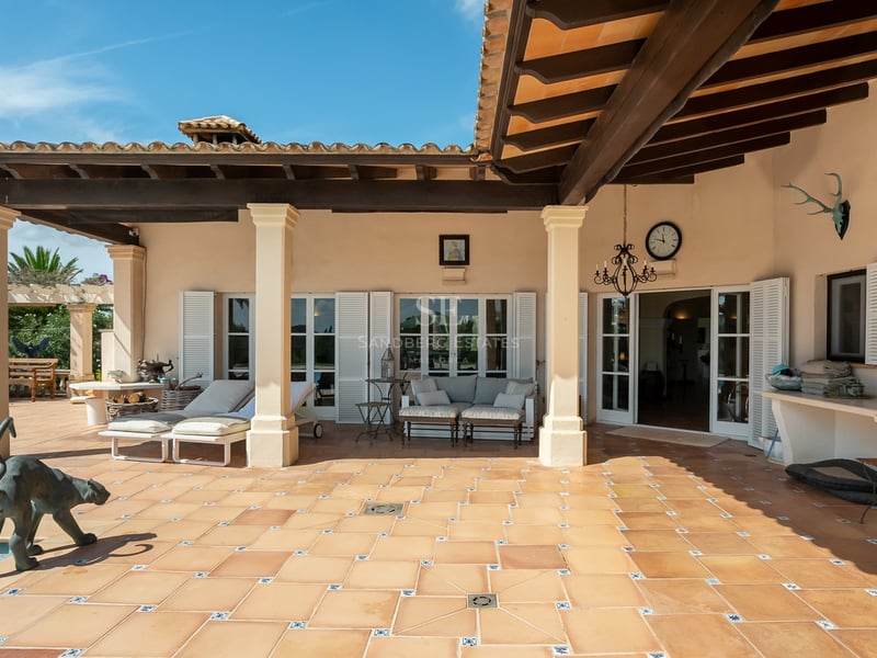 Terrasse en terre cuite avec poutres en bois sombre, piliers blancs, chaises longues et volets blancs sous le soleil.