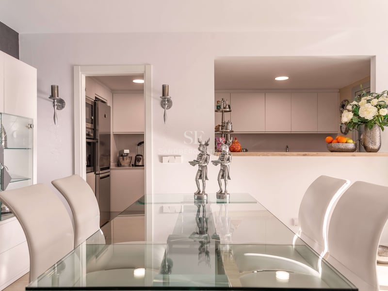 Zeitgenössisches Esszimmer mit Glastisch, weißen Stühlen und Blick in eine moderne weiße Küche durch eine Durchreiche.