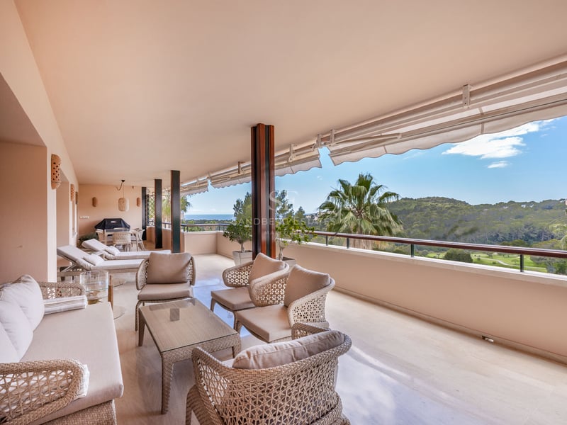 Grand balcon couvert avec mobilier en osier offrant une vue sur une vallée verdoyante et la mer par temps clair.