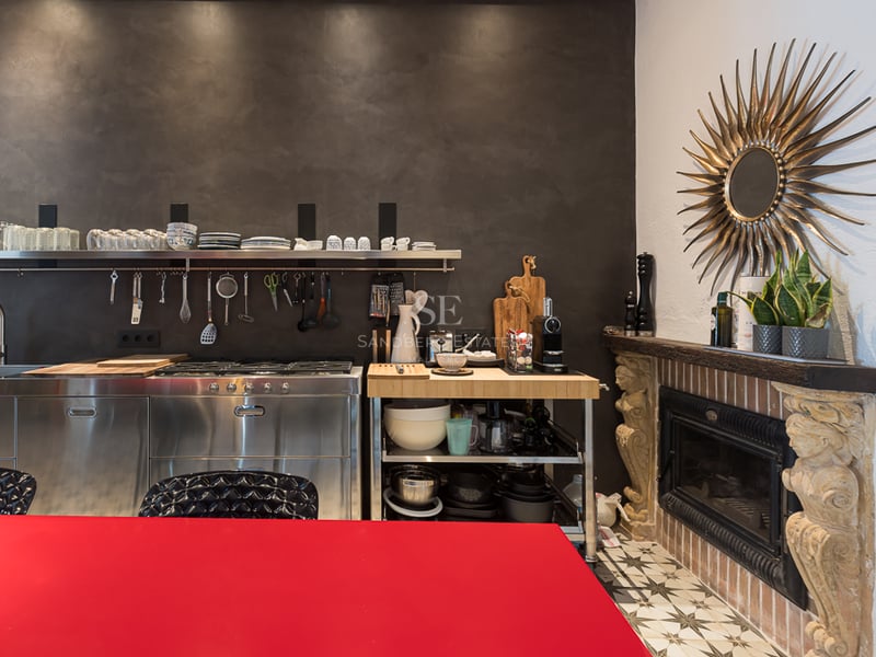 Cuisine moderne avec meubles en acier inoxydable, mur sombre texturé, table rouge et cheminée en pierre sculptée.