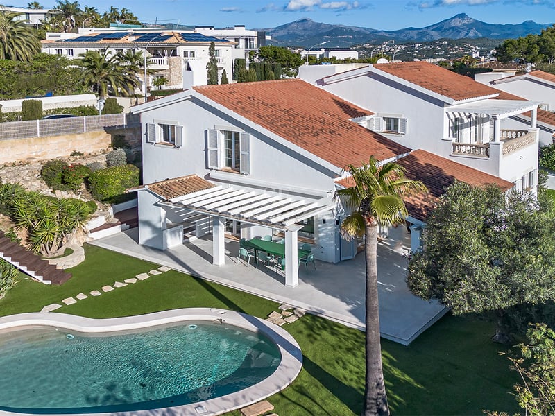 Flygfoto över en vit villa med terrakottatak, njurformad pool och trädgård mot en bakgrund av berg.