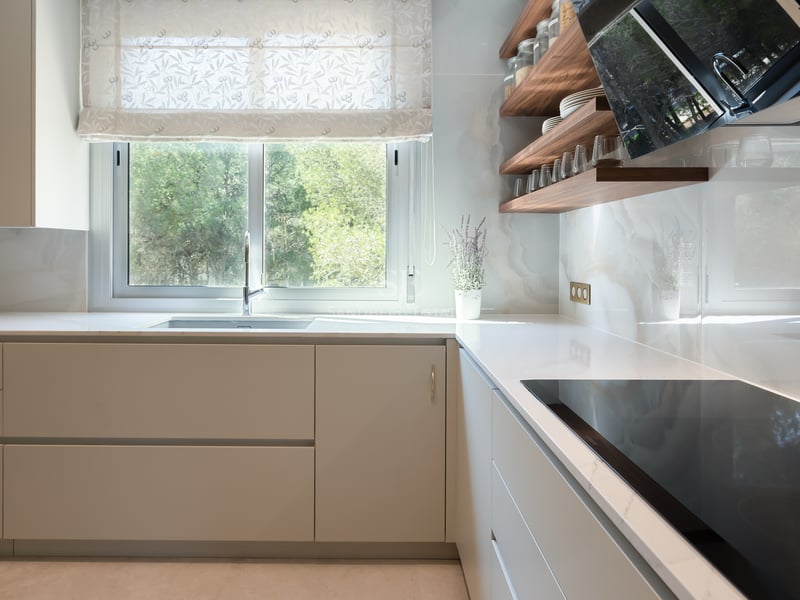 Minimalist off-white kitchen with marble-effect backsplash, wooden shelves, and a window overlooking lush greenery.