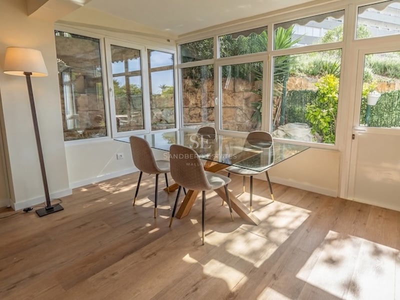 Comedor luminoso con mesa de cristal, sillas tapizadas en beige y amplios ventanales que dan a un jardín mediterráneo.
