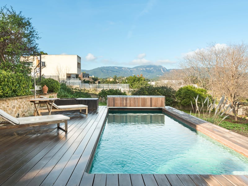 Rektangulär pool med mörkt trädäck, vita solstolar och panoramautsikt över bergen.