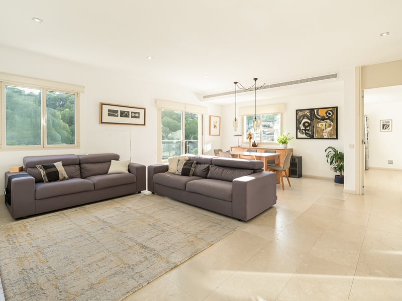 Rymligt och ljust vardagsrum med två grå soffor, ljusa klinkergolv och matplats i bakgrunden med stora fönster.