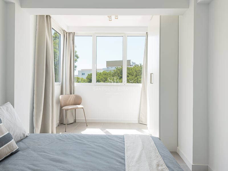 Une chambre lumineuse et moderne avec un lit gris, des placards intégrés blancs et une fenêtre donnant sur des arbres.