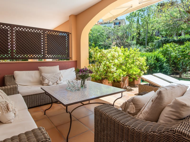 A covered terrace with wicker sofas, cream cushions, and a mosaic table overlooking a vibrant green garden.