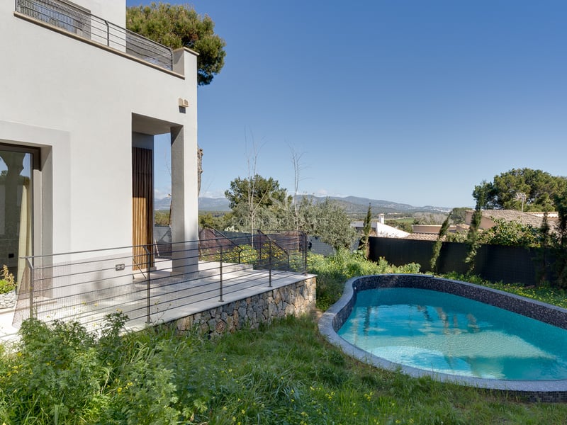 Modern vit villa med en kurvad blå mosaikpool, stenvägg och bergsutsikt under en klarblå himmel.