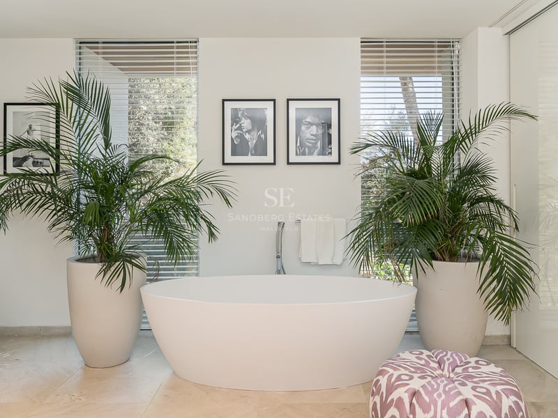 Freistehende weiße Badewanne flankiert von zwei großen Palmen vor bodentiefen Fenstern mit Jalousien.