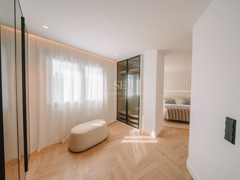 Dressing lumineux avec parquet en chêne, pouf beige et armoire vitrée menant à une chambre.