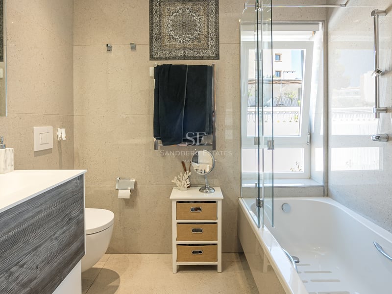 Modernt badrum med vitt badkar, glasskärm, kommod i träimitation och neutralt kakel med stenskänsla.