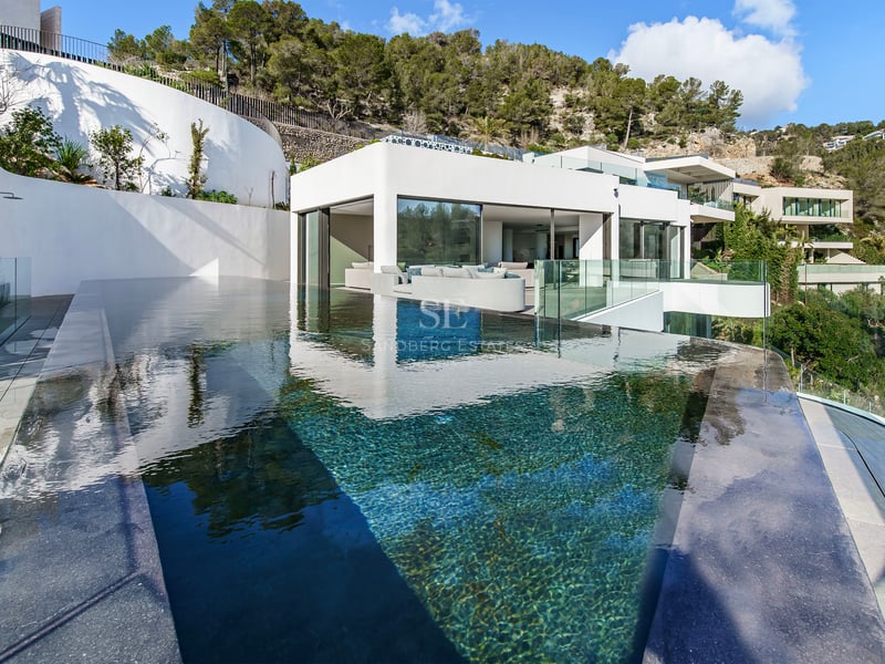 Piscine à débordement moderne aux eaux cristallines surplombant une villa blanche minimaliste nichée dans une colline.
