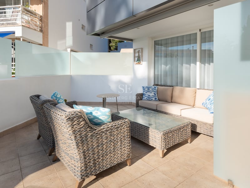 Terrasse moderne avec canapés en osier gris, coussins beige et bleus sur un patio ensoleillé avec parois en verre dépoli.