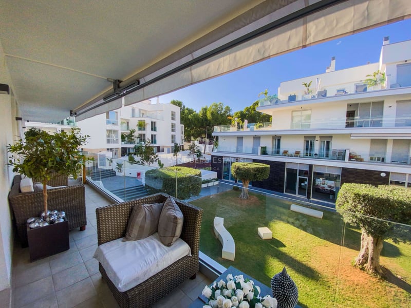 Terrasse mit Rattansesseln und Glasgeländer mit Blick auf einen Garten und moderne weiße Gebäude.