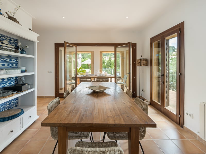 Intérieur avec grande table en bois, chaises en osier, sol en terre cuite et portes vitrées ouvrant sur une terrasse.