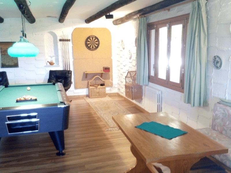 Sala de ocio con mesa de billar, diana, paredes de piedra blanca y vigas de madera oscura.