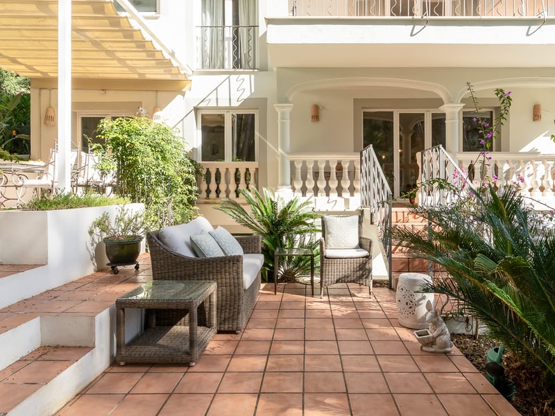 Sun-drenched terracotta tile terrace with wicker lounge furniture and lush greenery against a classic white villa facade.