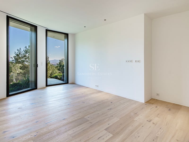 Une pièce vide avec du parquet en bois clair, des murs blancs et deux grandes baies vitrées donnant sur des arbres verts.